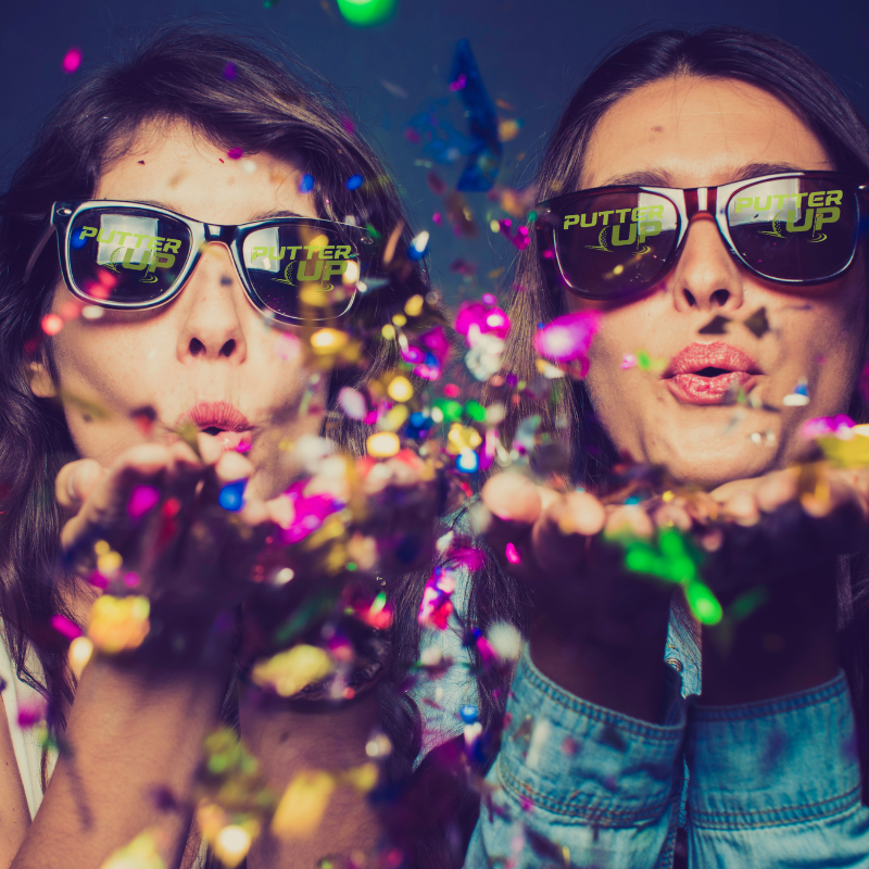 mini golf party at putter up two girls blowing confetti with sunglasses on that have the putter up logo on the lenses